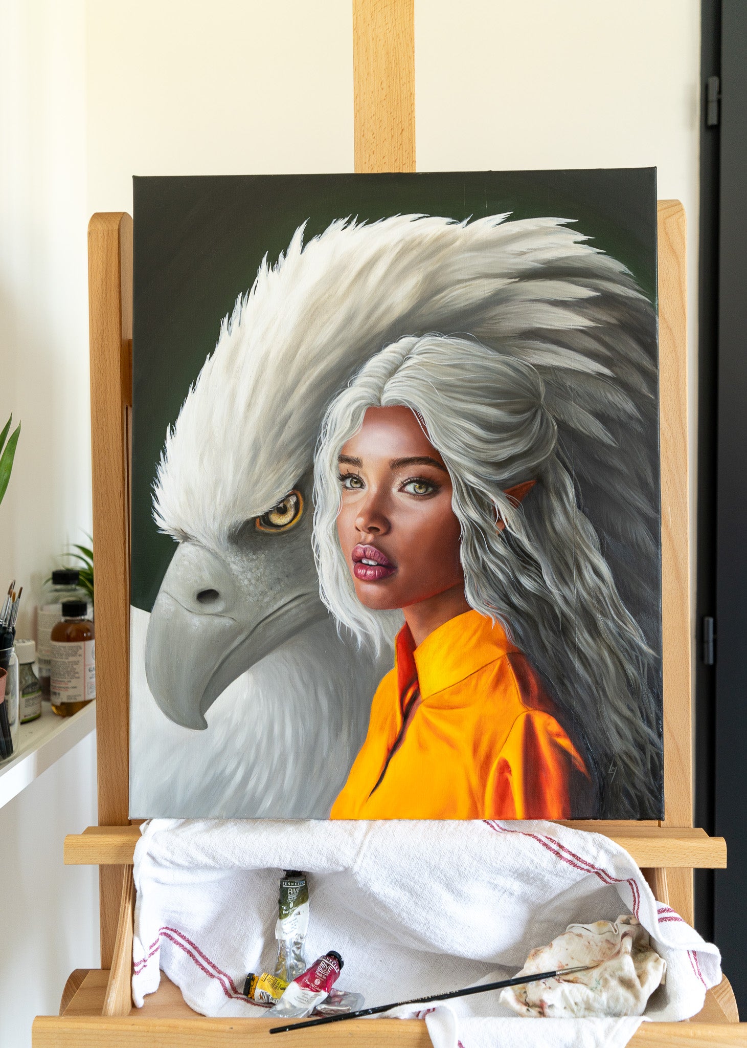 Le portrait d’elfe avec un aigle géant derrière, posé sur un chevalet, avec quelques tubes de peinture et un pinceau visible