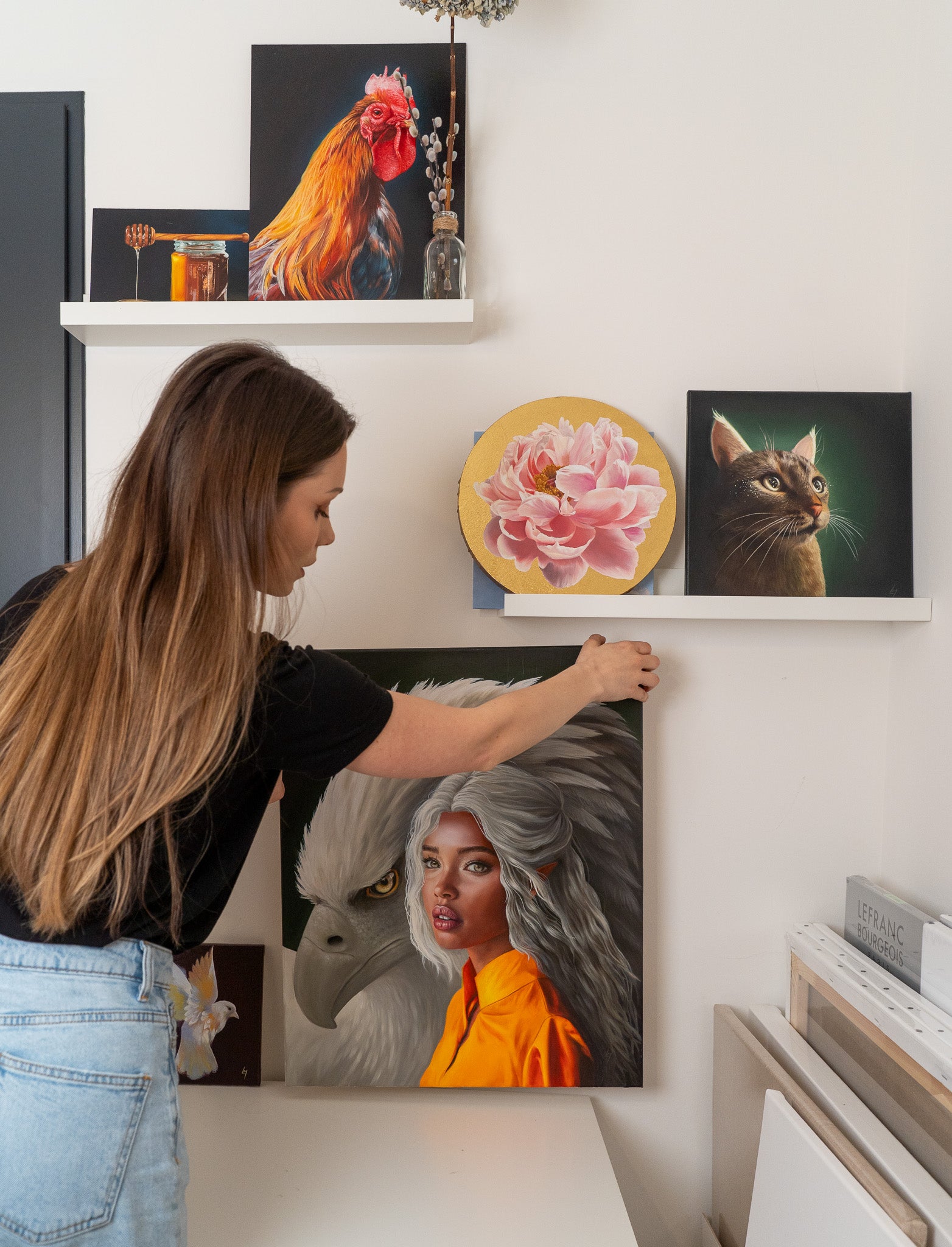 Dans son atelier, l’artiste présente un portrait d’elfe avec un aigle géant, avec d’autres peintures terminées visibles, telles qu’une fleur, un chat avec des galaxies dans les yeux, et un coq. À droite, des tableaux en cours d’attente